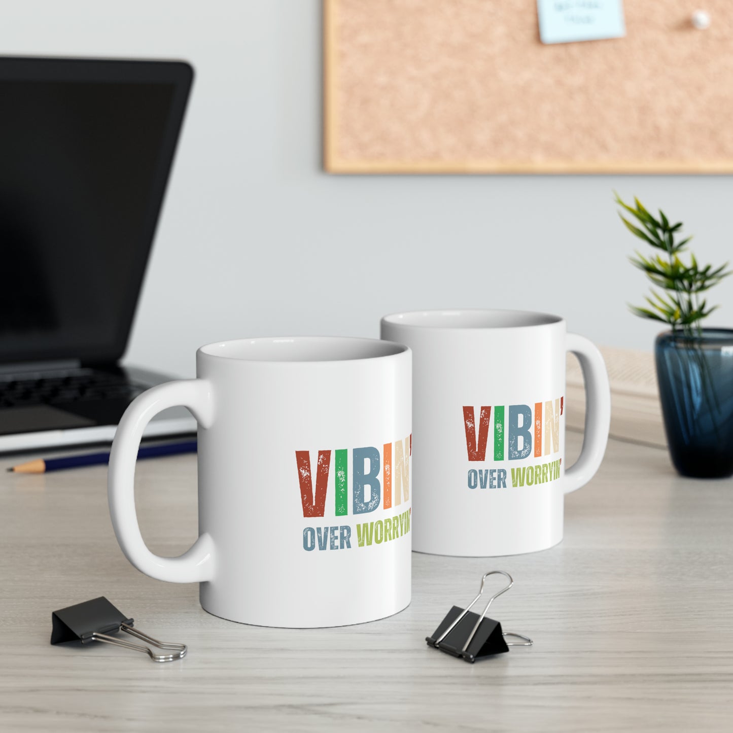 Two matching coffee mugs with the phrase "vibin' over worryin" printed on them, exuding an empowering and uplifting aura, sitting on a desk beside a laptop and a plant.