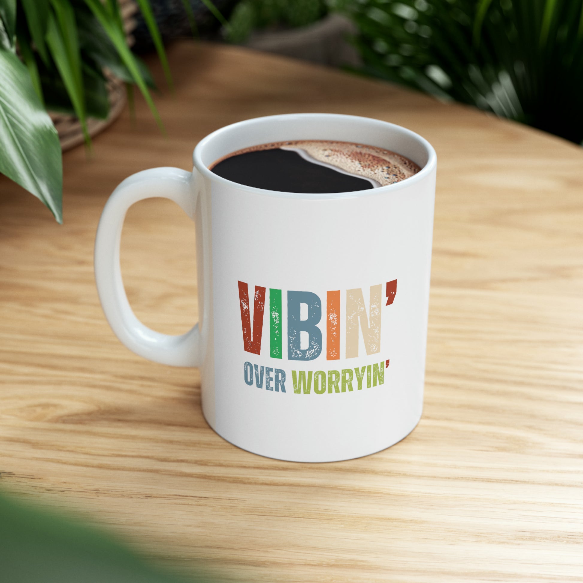 A white mug with the supportive inspirational quote "vibin' over worryin'" printed on it sits on a wooden surface, with a lush green plant in the background. The mug is filled with coffee.