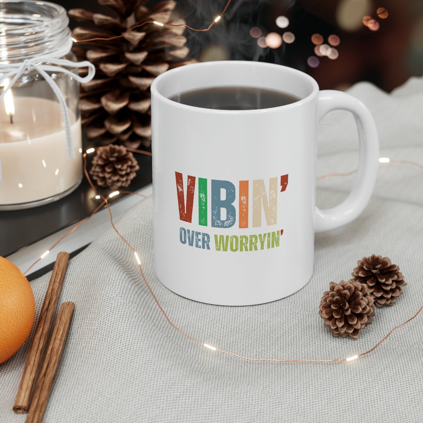 A cozy and uplifting moment with a Vibin' Over Worryin' Ceramic Mug 11oz amidst a festive setting with pine cones, an orange, cinnamon stick, and.