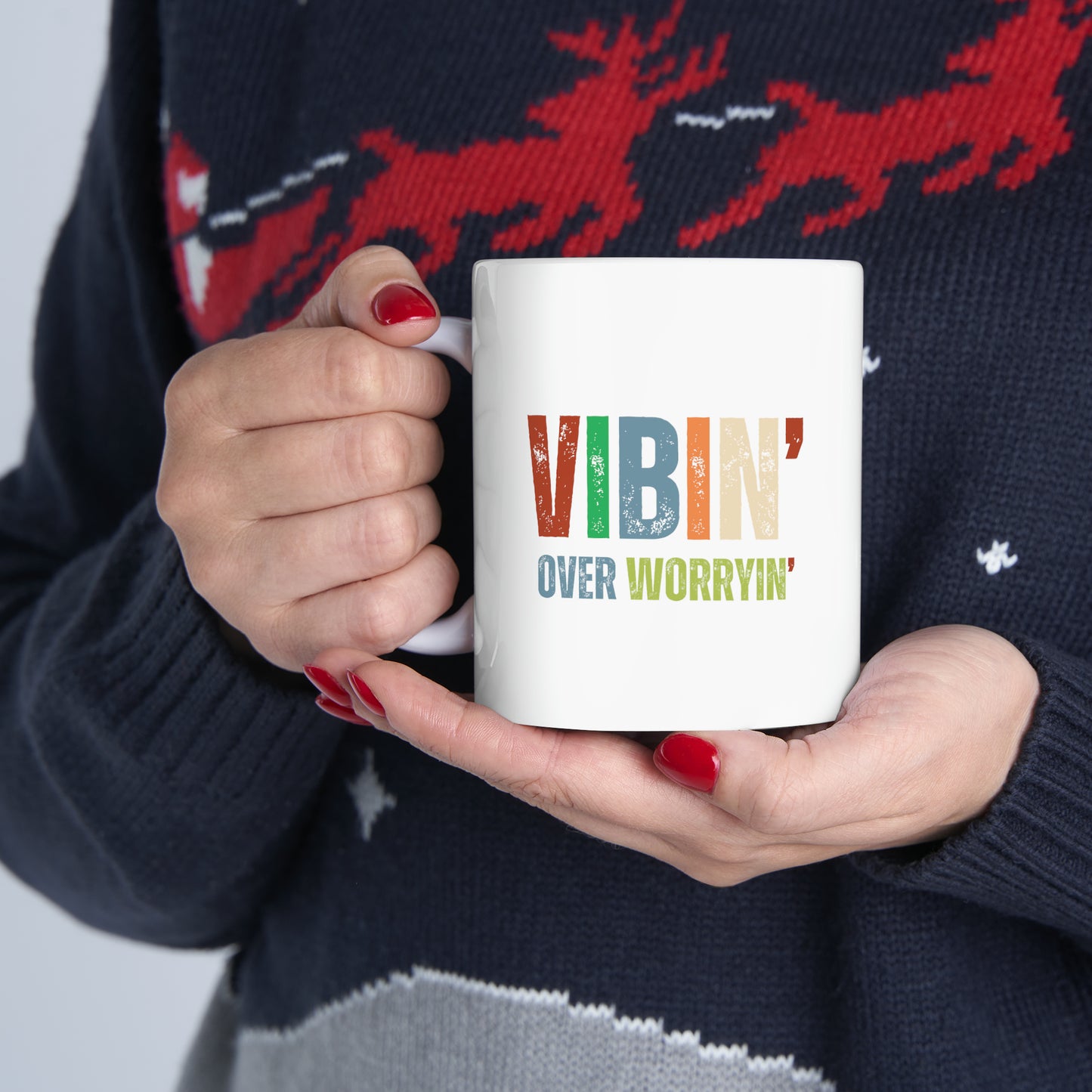 A person wearing a cozy sweater holds a white mug with the inspirational message "vibin' over worryin'" printed in colorful letters.