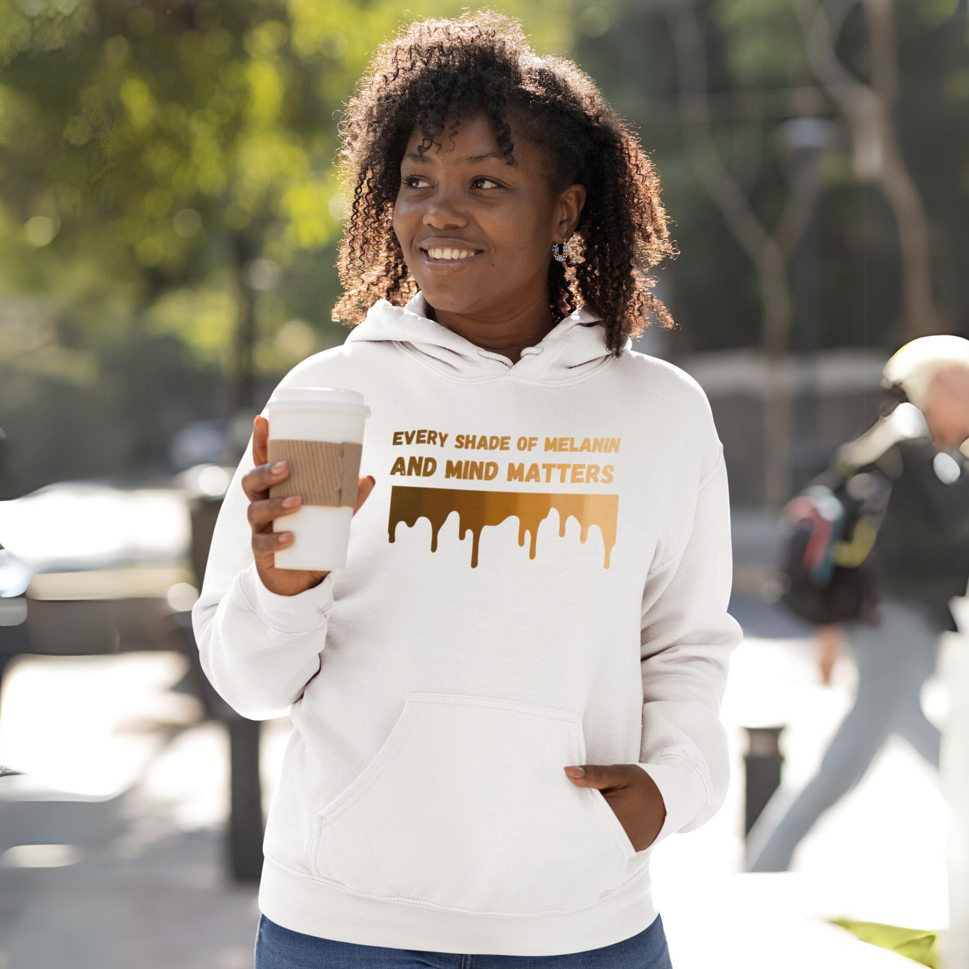 A woman smiling while holding a coffee cup, wearing the Every Shade Of Melanin Unisex Hooded Sweatshirt with the message "every shade of melanin and mind matters" printed on it, celebrating diversity and mental health awareness in an inspirational way.