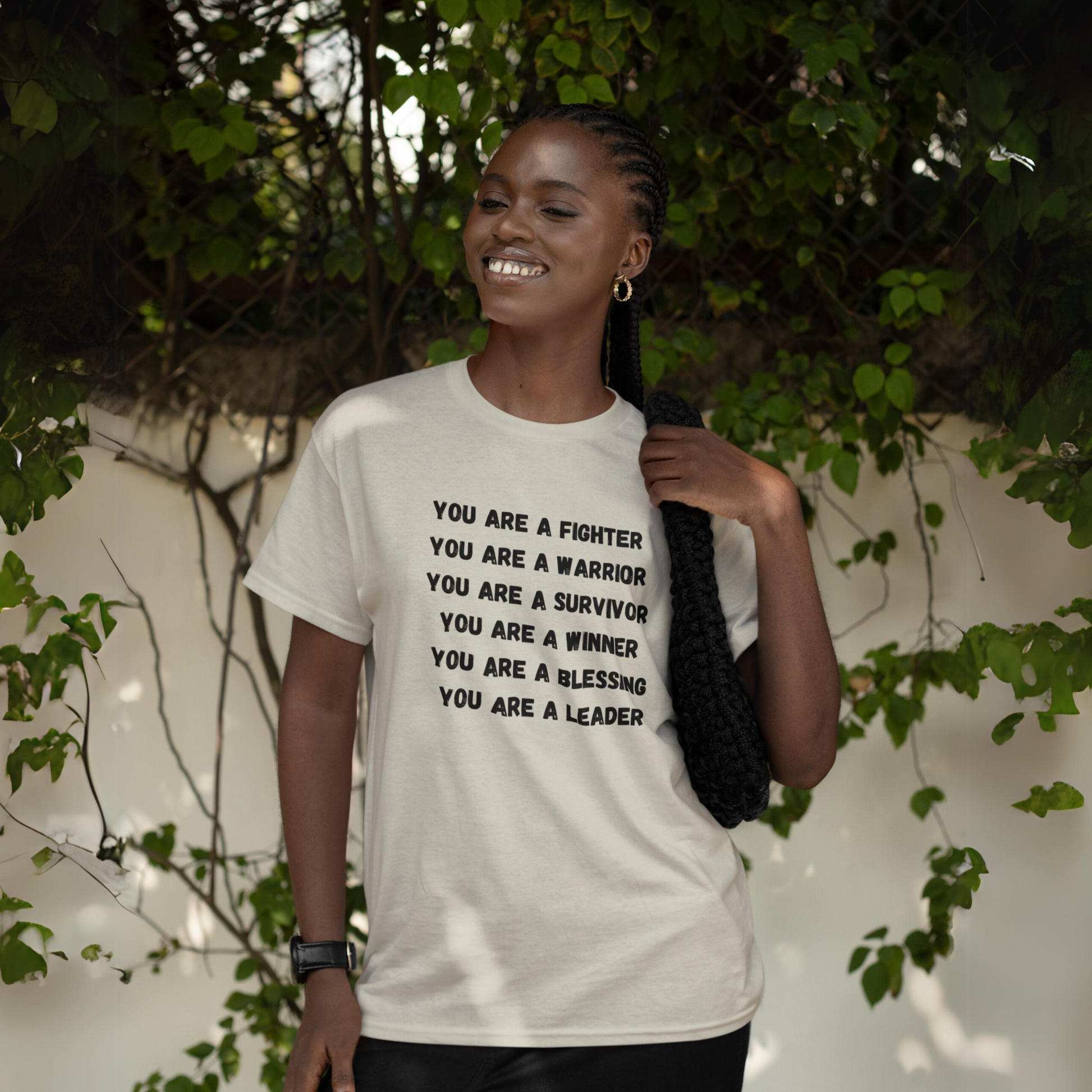 A smiling woman confidently wearing a "You Are a Fighter" unisex heavy cotton t-shirt among green foliage.