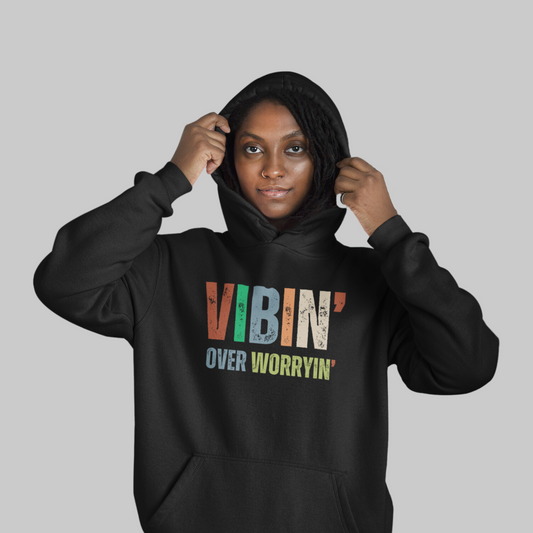 A confident woman wearing a Vibin' Over Worryin' Unisex Hooded Sweatshirt with the inspirational phrase "vibin' over worryin'" printed on the front.