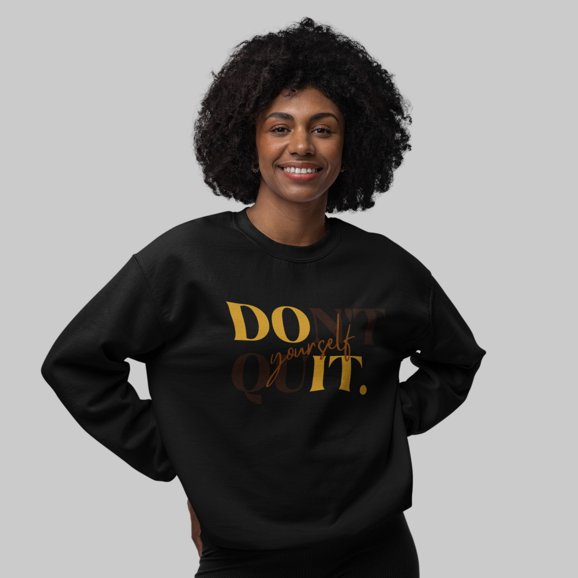 A smiling woman with curly hair wearing a black DOn't quIT Yourself Unisex Crewneck Sweatshirt featuring the supportive quote "don't quit your daydream" in gold letters for mental health awareness.