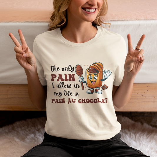 A woman sits on the floor in front of a bed, smiling and making peace signs with both hands. She wears "The Only Pain I Allow In My Life - Chocolat" unisex jersey short sleeve tee, which is cream-colored and features a cartoon croissant holding a chocolate bar and spoon with the text, "The only pain I allow in my life is pain au chocolat.