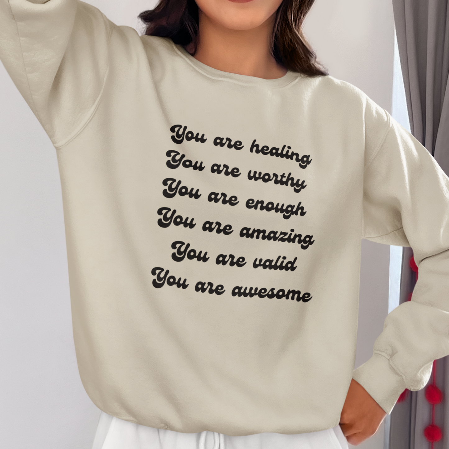 A person wearing a You Are Healing Unisex Crewneck Sweatshirt with an uplifting empowering message printed on it: "you are healing, you are worthy, you are enough, you are amazing, you are valid, you are awesome".