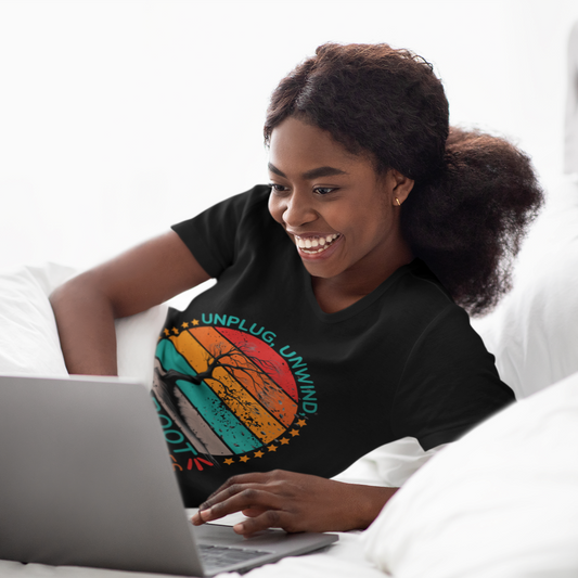 A cheerful young woman with a radiant smile lies on a white bed while using a laptop. She is wearing a black t-shirt with a colorful graphic that reads 'UNPLUG UNWIND UPROOT the bad vibes' alongside an artistic depiction of a tree with a vibrant sunset in the background, representing different mental states.
