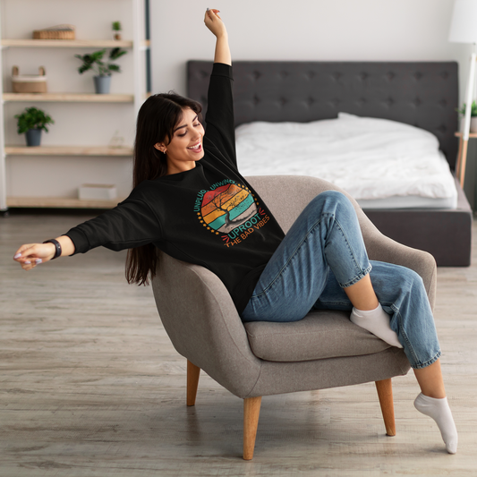 A relaxed woman with long hair stretches joyfully in a modern armchair, wearing a casual black sweatshirt with a colorful, circular design that reads 'UNPLUG UNWIND UPROOT THE BAD VIBES.' She's paired it with comfortable blue jeans and white socks, sitting in a well-lit room with a cozy bed in the background, embodying a sense of comfort and ease.