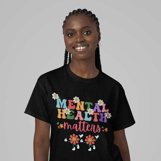 A woman with braided hair smiles at the camera, wearing a black "Mental Health Matters Unisex Softstyle T-Shirt." The shirt features colorful text that says "Mental Health Matters," surrounded by decorative flowers and two smiling flower characters at the bottom. The background is plain gray. Show your support proudly with this stylish tee!