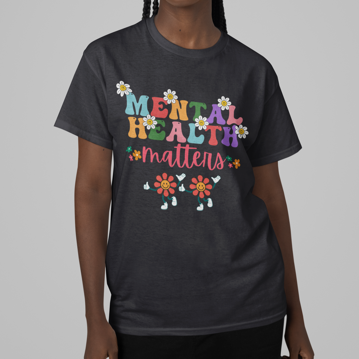 A woman with braided hair smiles at the camera, wearing a black "Mental Health Matters Unisex Softstyle T-Shirt." The shirt features colorful text that says "Mental Health Matters," surrounded by decorative flowers and two smiling flower characters at the bottom. The background is plain gray. Show your support proudly with this stylish tee!